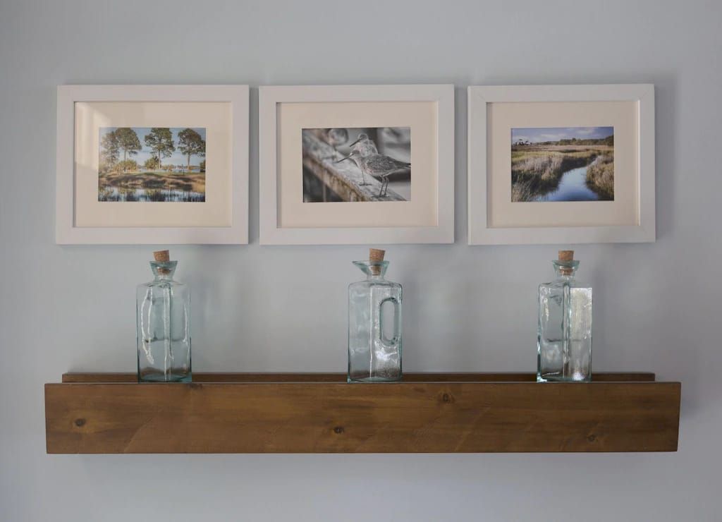 Three pictures are hanging on a wall above a wooden shelf with three bottles on it.