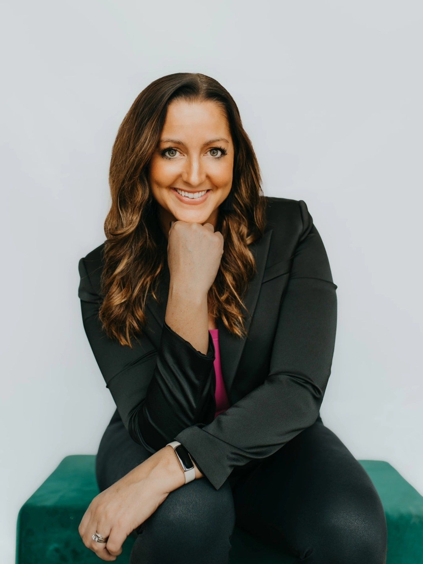 Lisa in black blazer smiles, resting chin on hand. Sitting on a green bench, against a white backdrop.