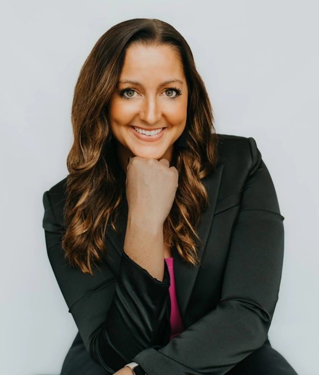 Lisa in a black blazer smiles, resting her chin on her hand, against a white backdrop.
