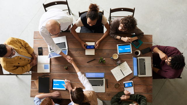 People at a wooden table in a meeting, one handshake. Laptops, tablets, coffee, and notebooks are visible.