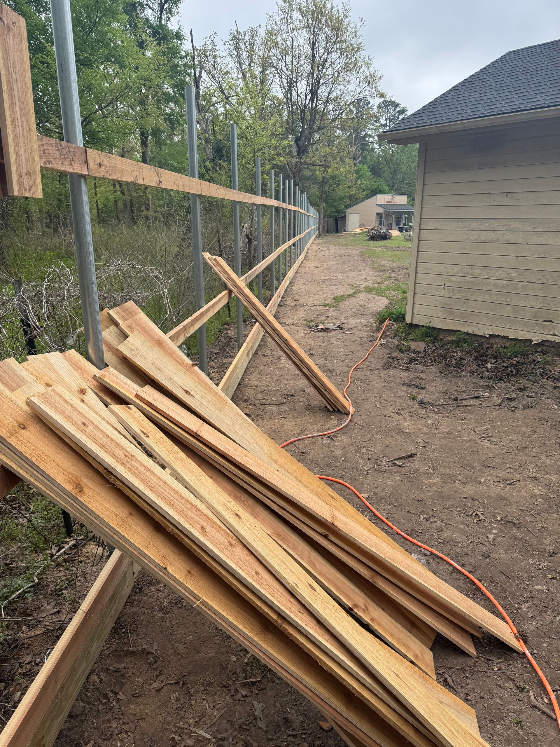 Stacked wooden boards beside a fence and beige building on a dirt path outdoors