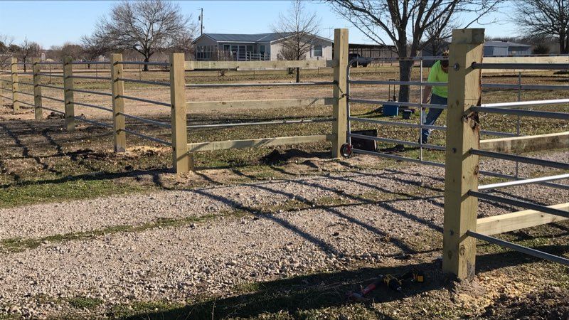 Gravel farm entrance with wooden fence and gate opening to a grassy field and barn in the distance