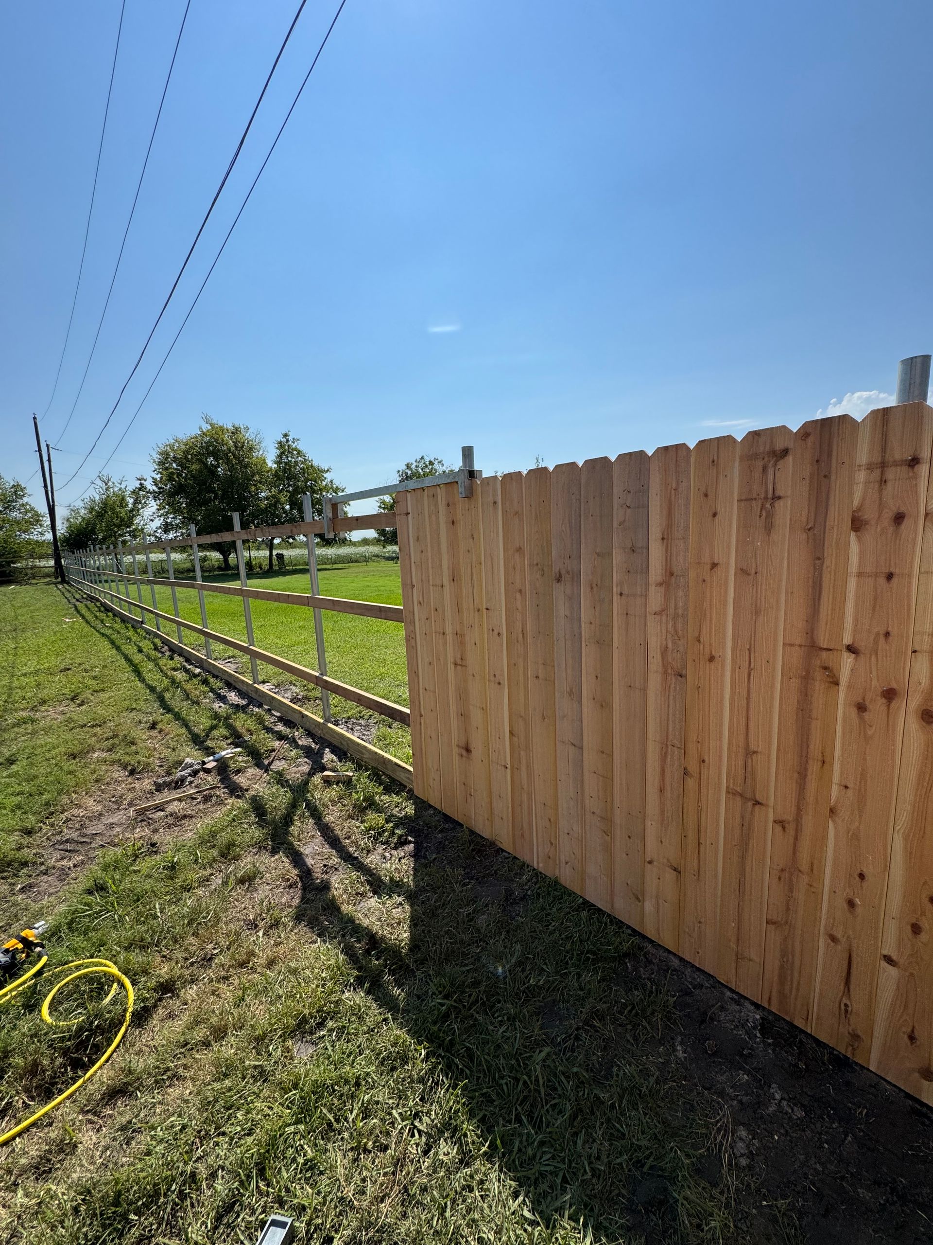 Wooden fence beside a grassy field under a clear blue sky, with utility lines overhead.