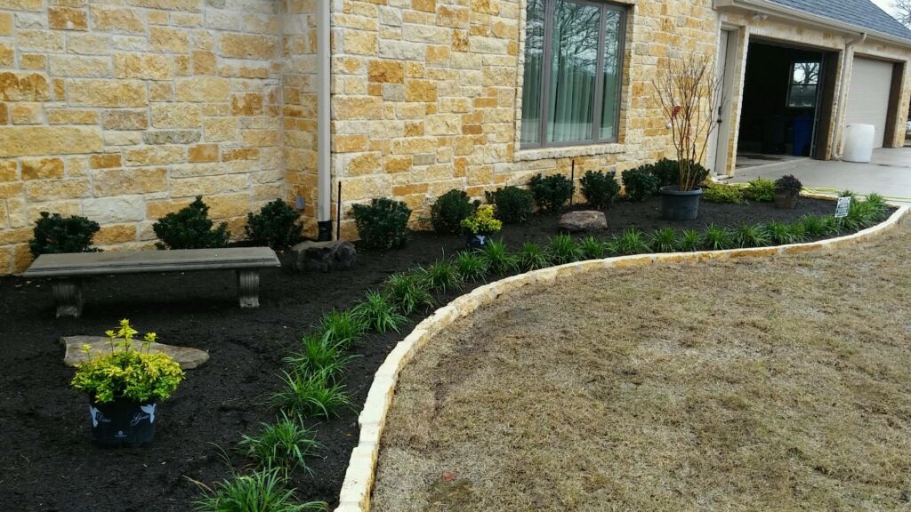 Stone house garden bed with black mulch, shrubs, a bench, and a curved white border beside a dry lawn.