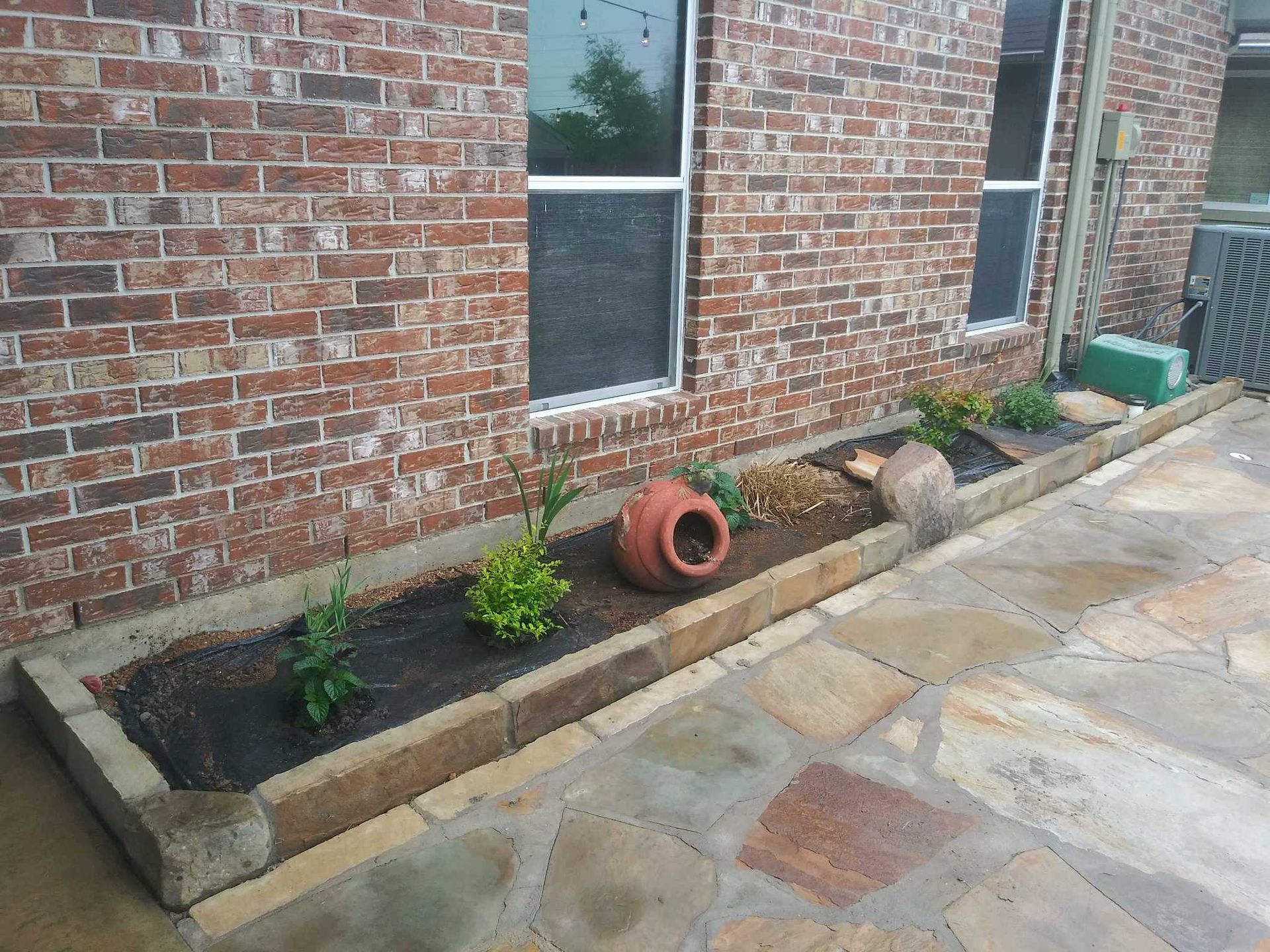 Brick building with a narrow raised garden bed, small plants, and a tipped terracotta pot along a stone walkway.