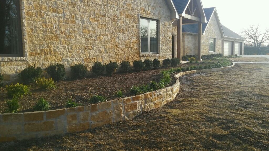 Stone-faced house with curved brick planter bed and shrubs beside a gravel driveway
