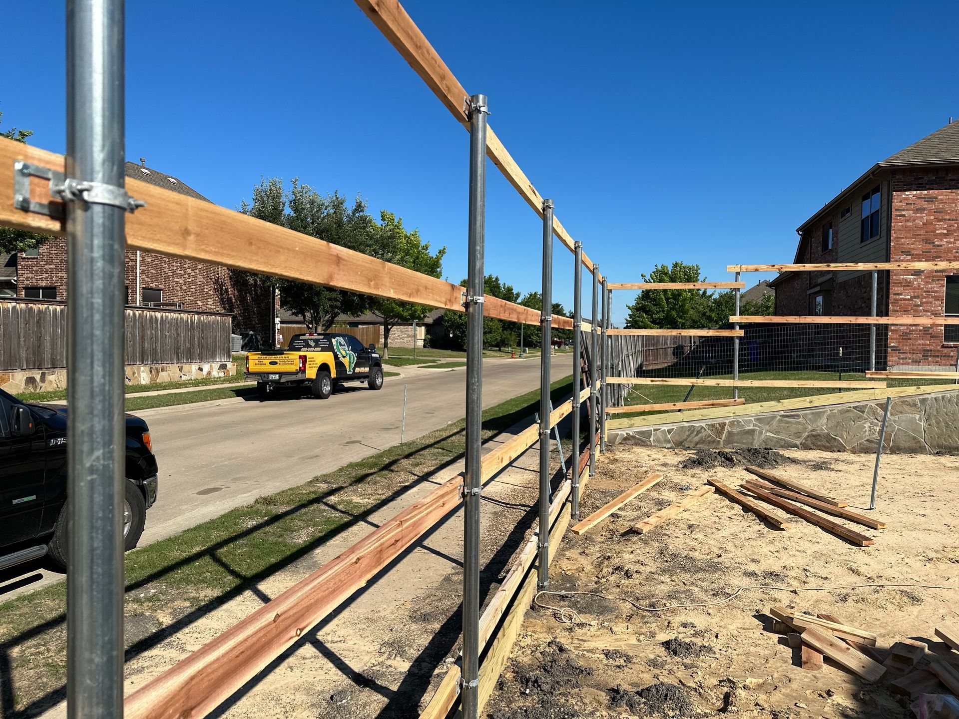 Construction site with metal fence framing a dirt area, brick buildings, and a yellow utility truck under a blue sky