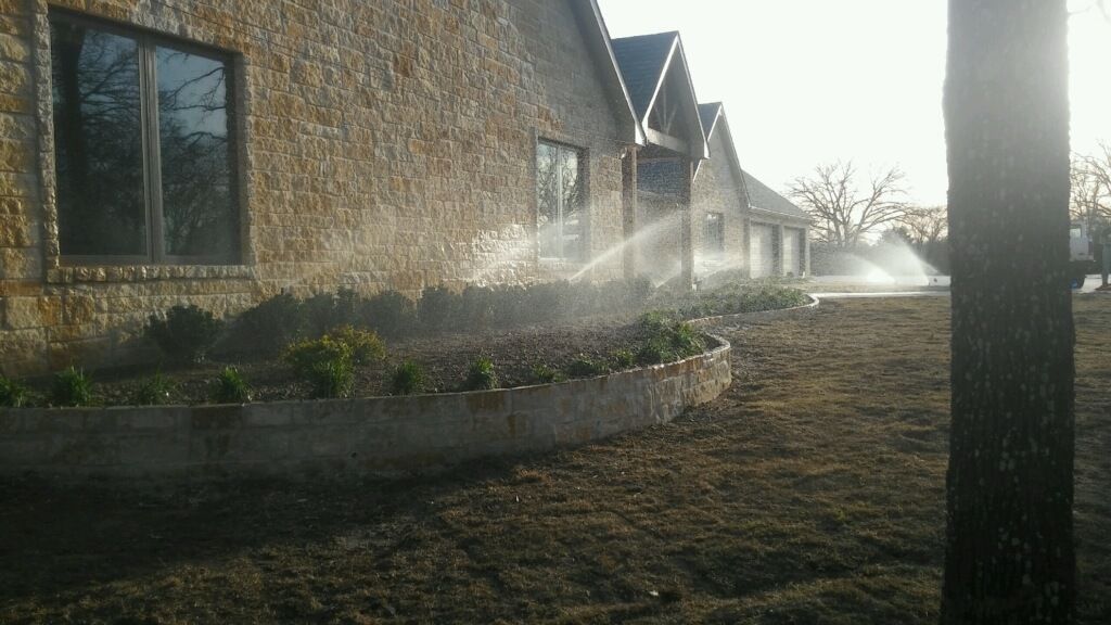 Sprinklers watering a landscaped yard beside a stone house at sunrise.