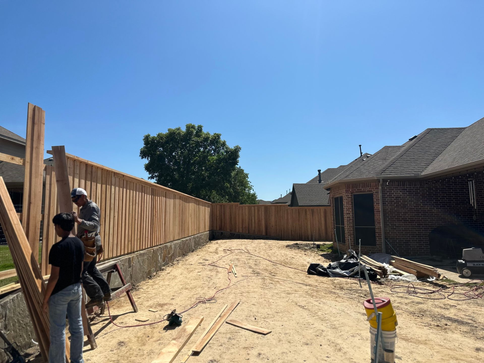 Backyard under construction with workers, wooden fence, dirt ground, and a house on the right under a blue sky