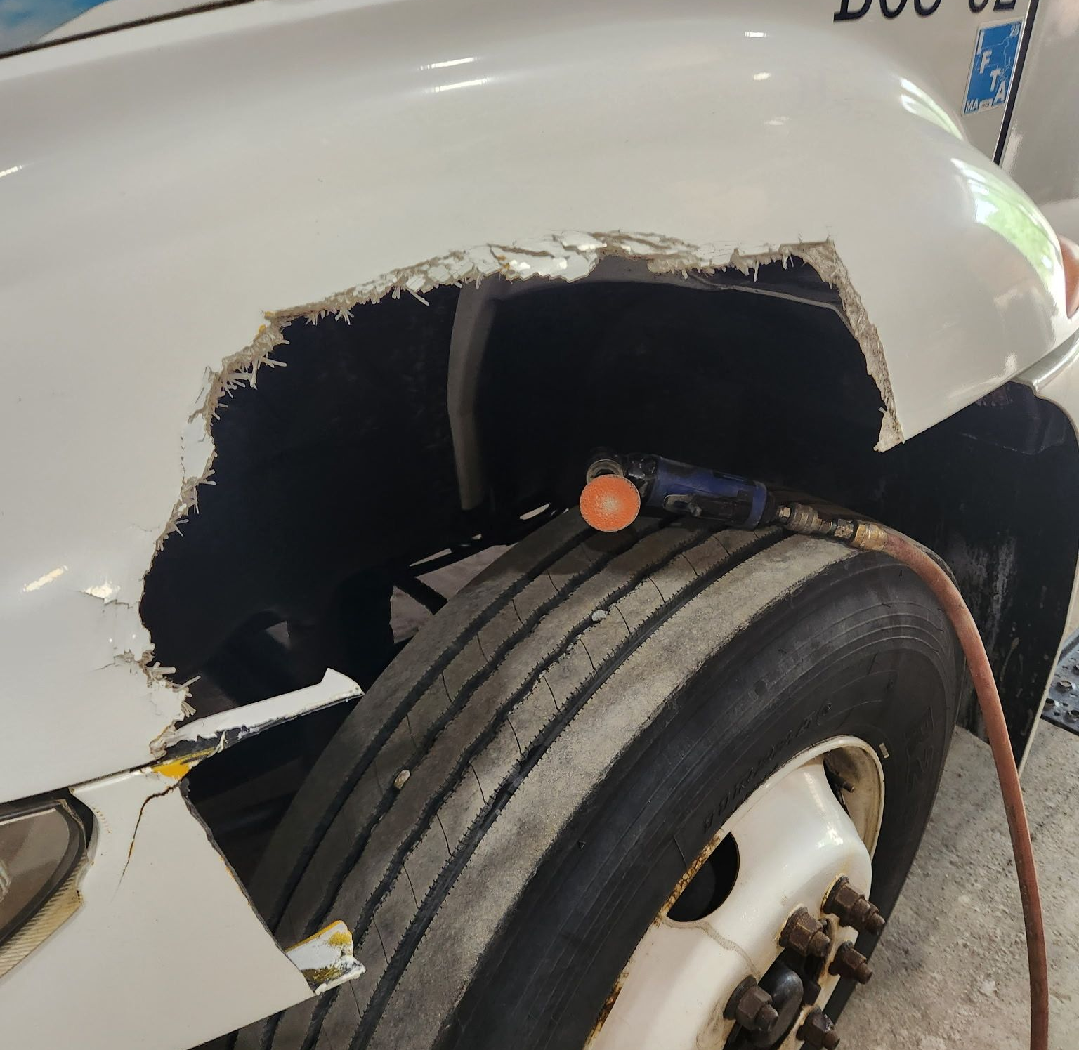 Damaged white truck fender with a large hole near the tire, showing the wheel well. An air hose is attached.