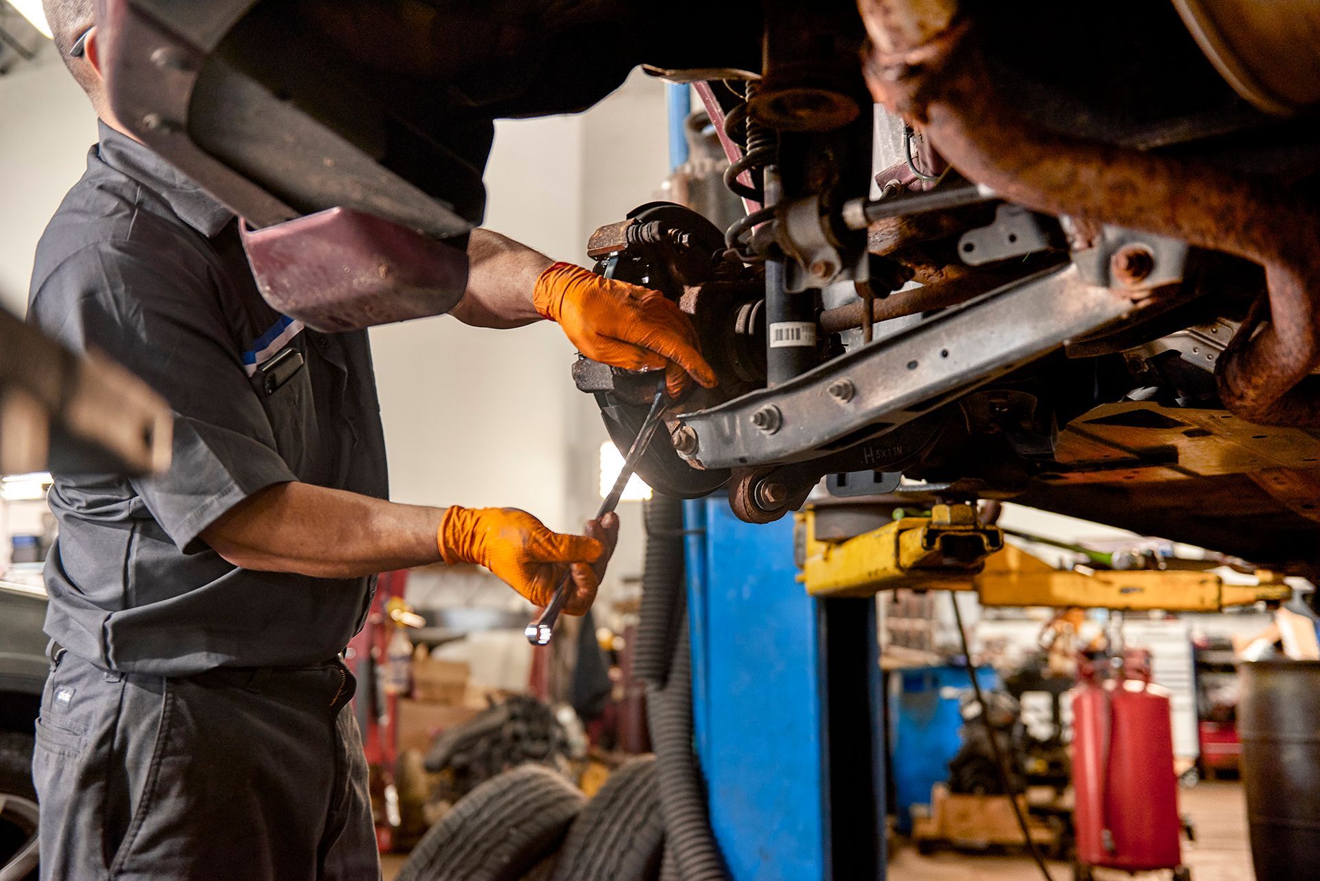 Mechanic in gray shirt and orange gloves working under a car on a lift in an auto repair shop