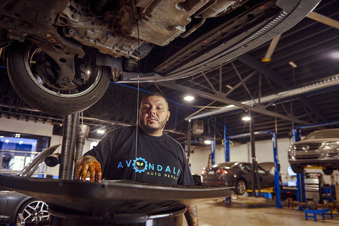Mechanic standing beneath a car on a lift in an auto repair shop, with tools and another car in the background.