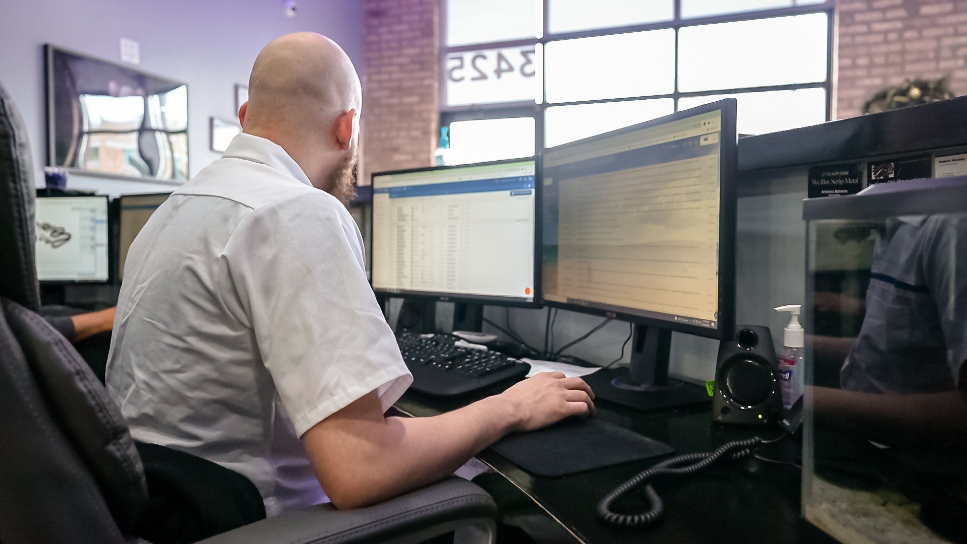 Person working at a desk with dual monitors in an office, facing a bright window.