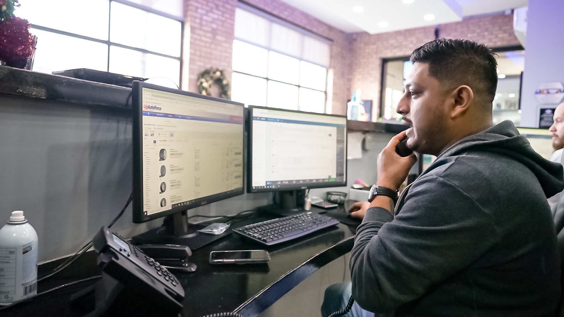 Person on a phone at a desk with dual monitors in an office, typing and looking at the screens.