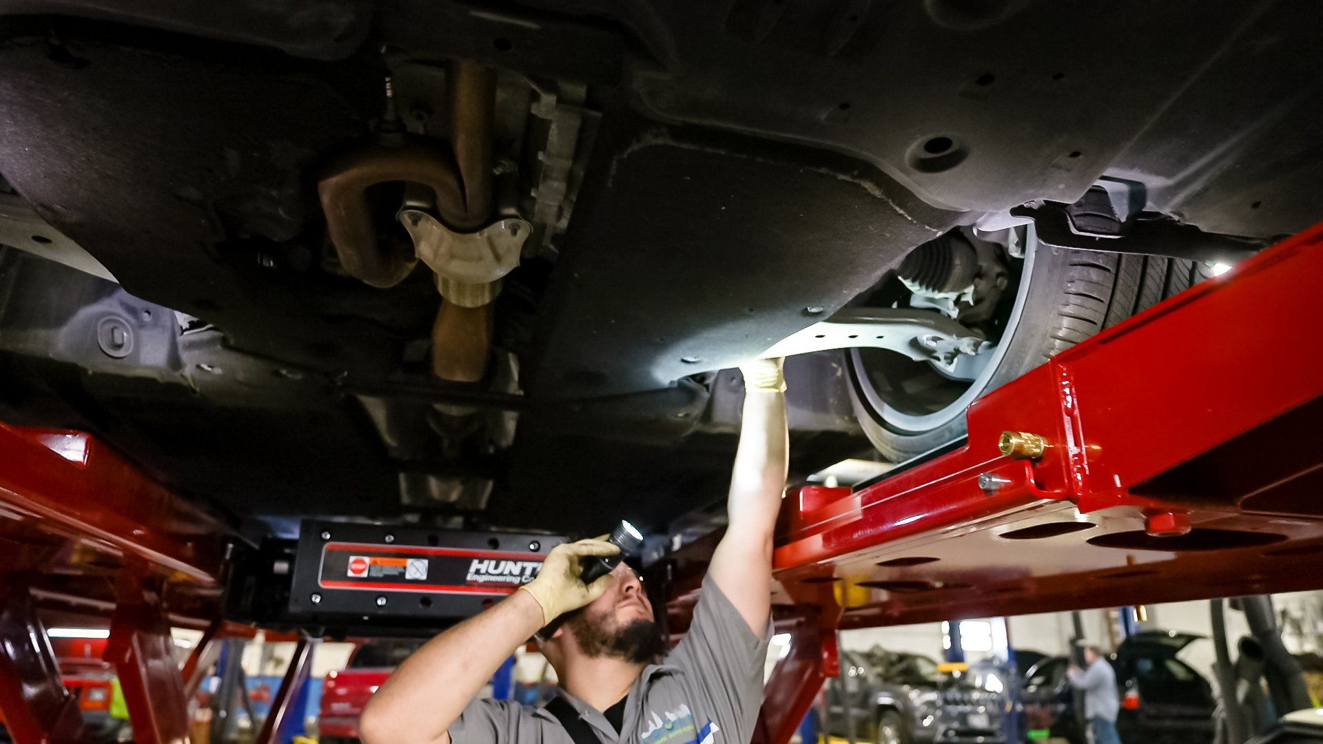 Mechanic under a lifted car in a red auto shop, reaching up to inspect the undercarriage