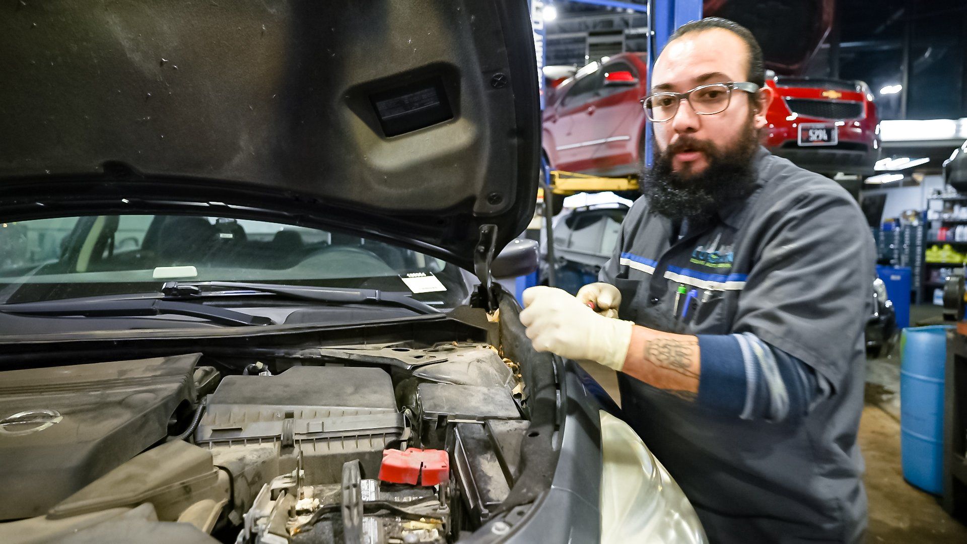 Mechanic beside an open car hood in an auto shop, inspecting the engine with gloves on.