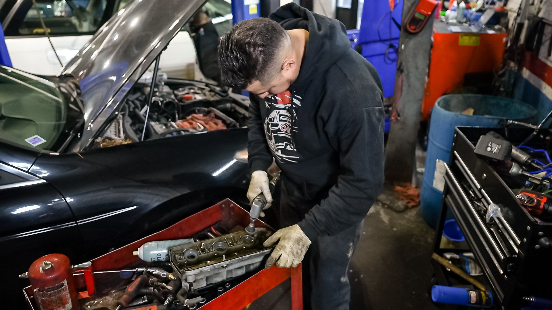 Mechanic working under the hood of a black car in a busy auto repair shop