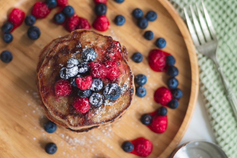 Pila di pancake ricoperti di lamponi, mirtilli e zucchero a velo, su un piatto di legno.