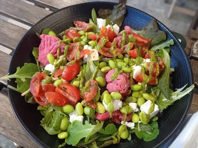 Insalata con foglie verdi, pomodori, edamame, cavolfiore rosa, formaggio feta e condimento verde in una ciotola nera.