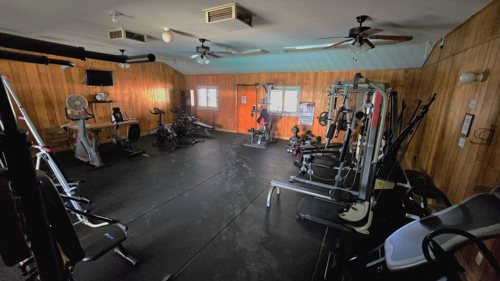 Gym room with exercise machines, black flooring, wood-paneled walls, and an orange back wall near a window.