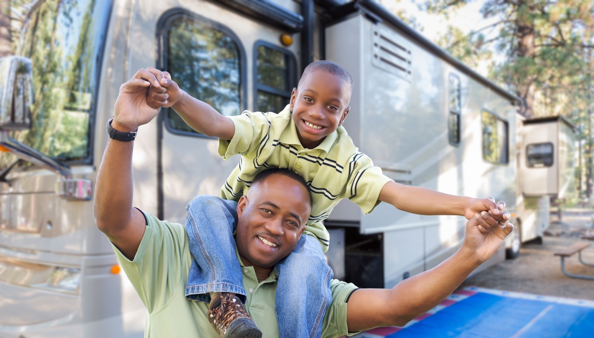 Person carrying a child on their shoulders beside a parked RV at a wooded campground.