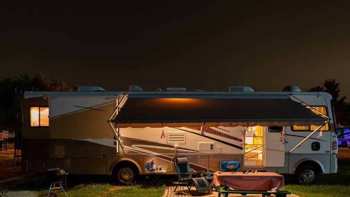 RV parked at a campsite at night with awning extended, outdoor chairs, table, and lights. RV parked at a campsite at night with awning extended, outdoor chairs, table, and lights.