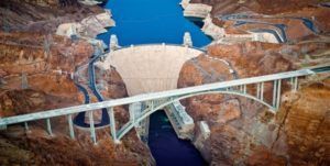 Aerial view of the Hoover Dam with a large concrete arch bridge in the foreground. Blue water and brown rock formations surround the dam. Aerial view of Hoover Dam and the Mike O'Callaghan–Pat Tillman Memorial Bridge. The concrete dam is flanked by brown rock formations.