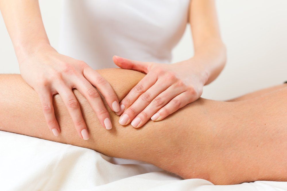 Hands massaging a person's leg lying on a white surface; in a light-filled setting.