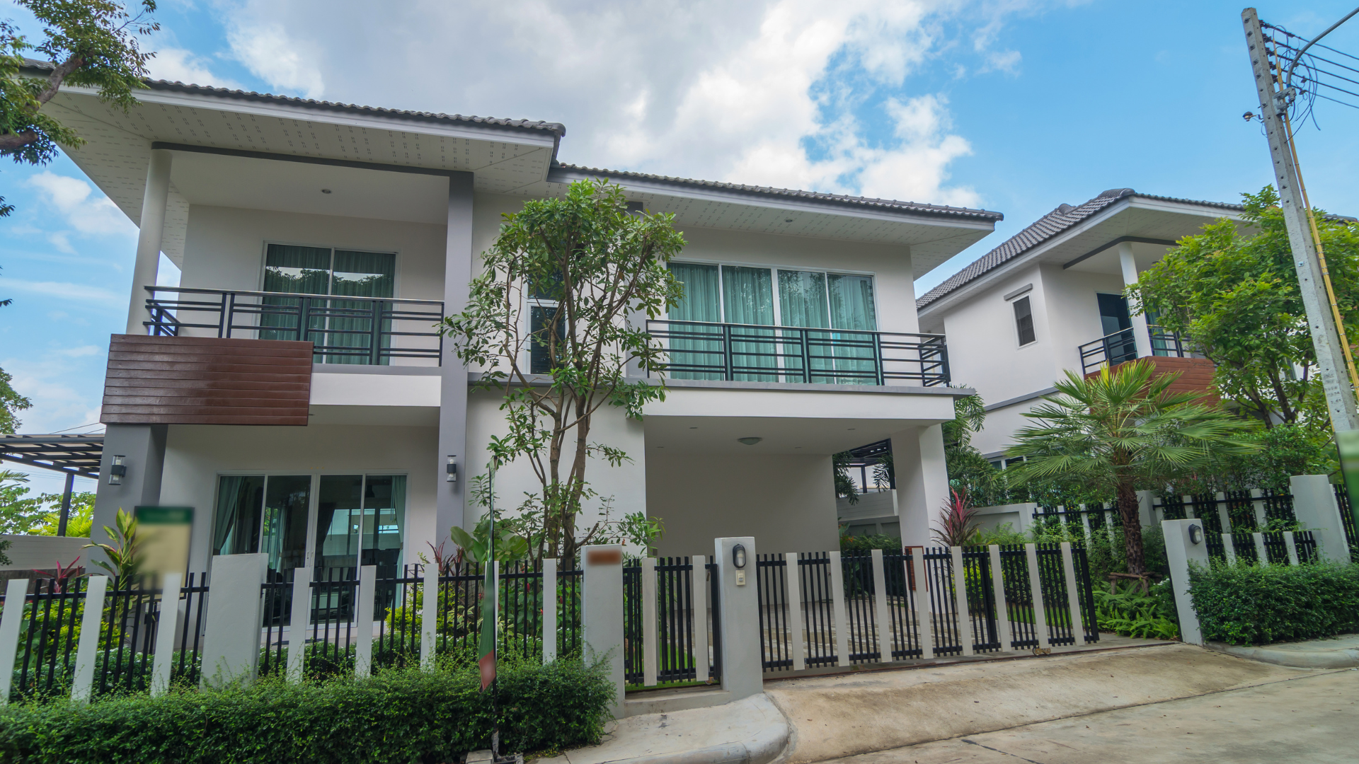 A two-story white residential house with a balcony, dark trim, and a modern gated fence under a blue, cloudy sky.