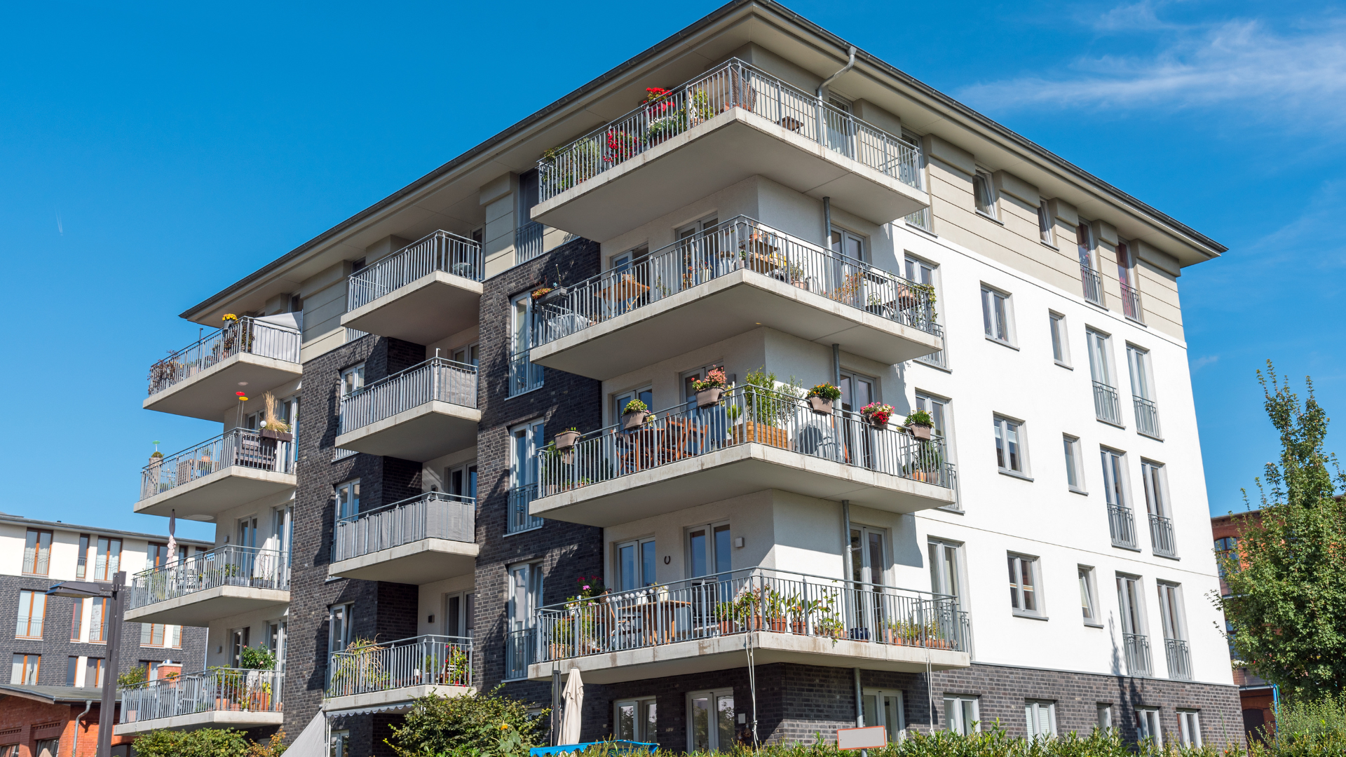 A modern, multi-story apartment building with balconies and white and dark grey exterior walls under a clear blue sky.