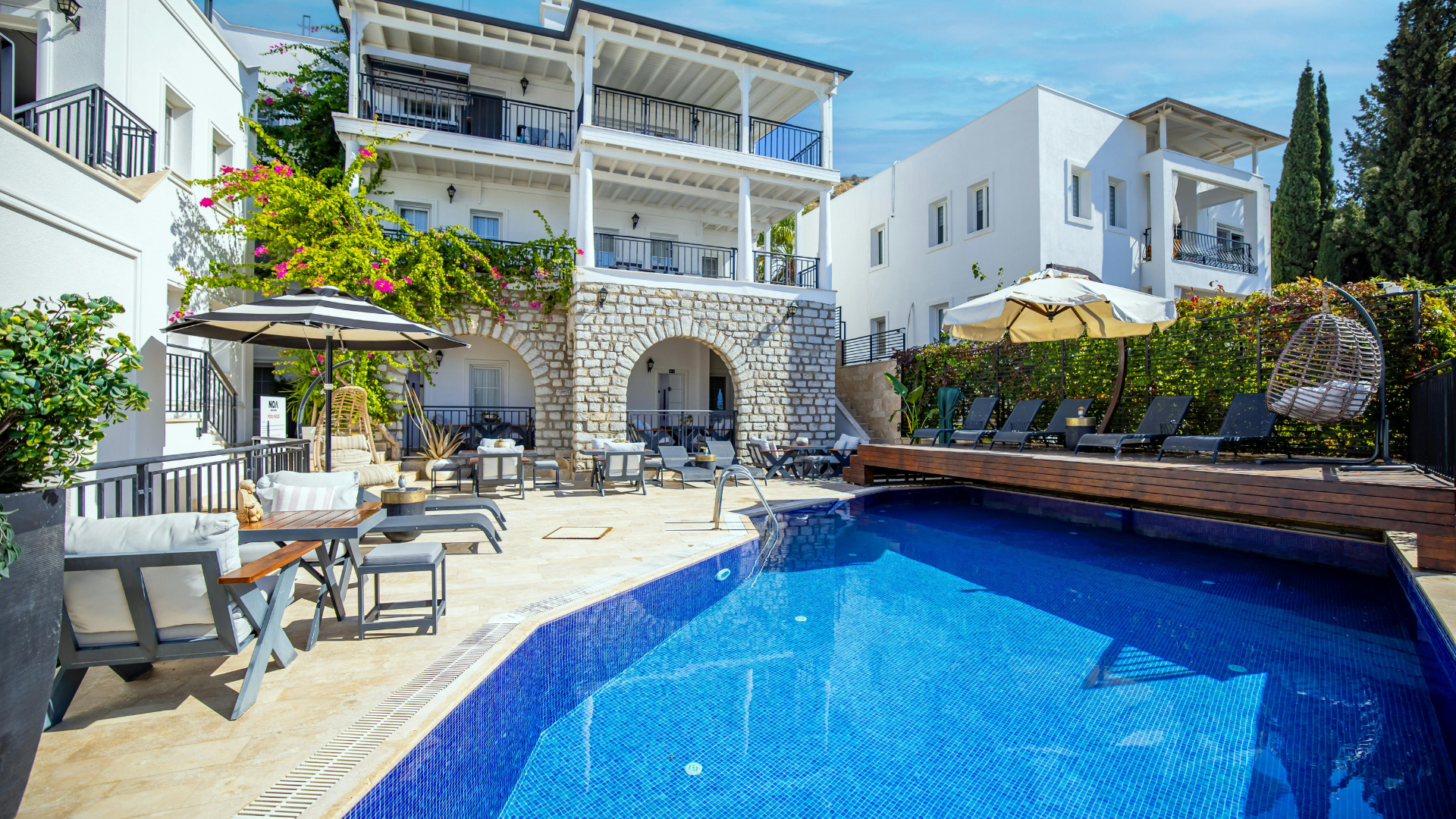 A luxurious swimming pool area with white villas, stone archways, lounge chairs, and umbrellas under a bright blue sky.