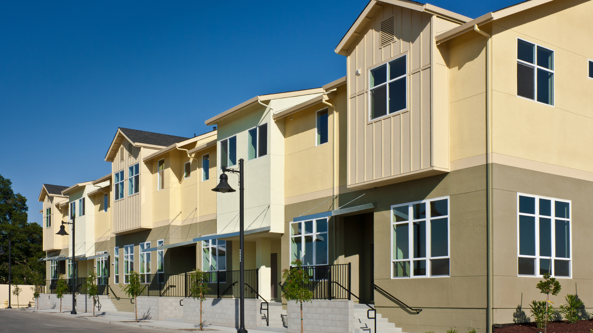 A row of modern townhomes with tan and beige siding, vertical accents, and large windows under a clear blue sky.