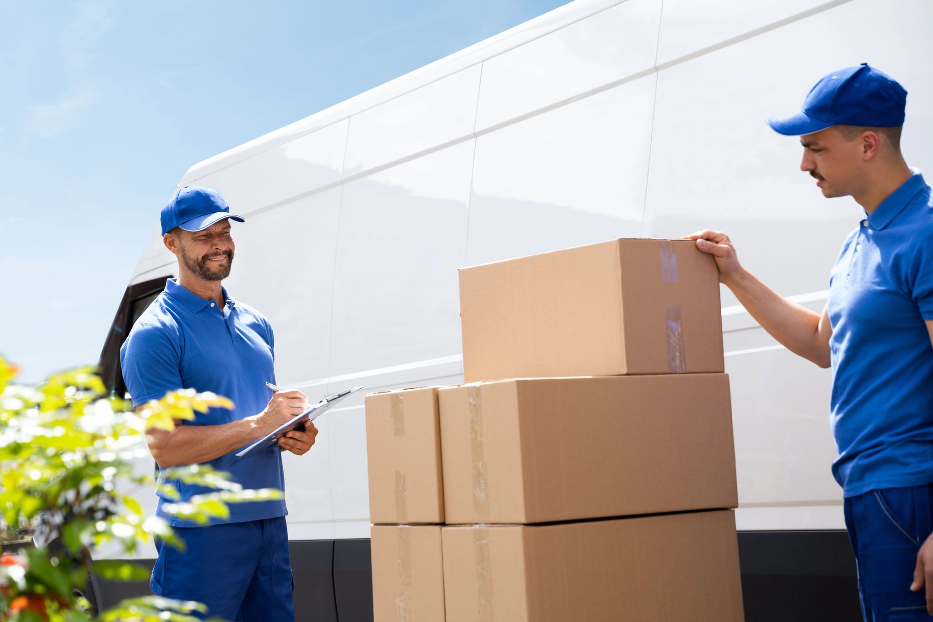 Two movers in blue uniforms unloading cardboard boxes from white moving truck outdoors. Two movers in blue uniforms unloading cardboard boxes from white moving truck outdoors.