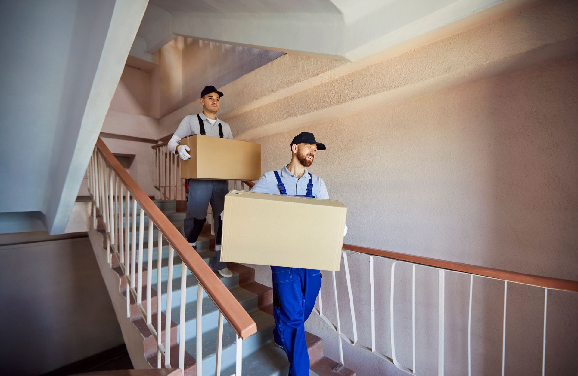 Two movers carrying large cardboard boxes down a staircase in an apartment building.