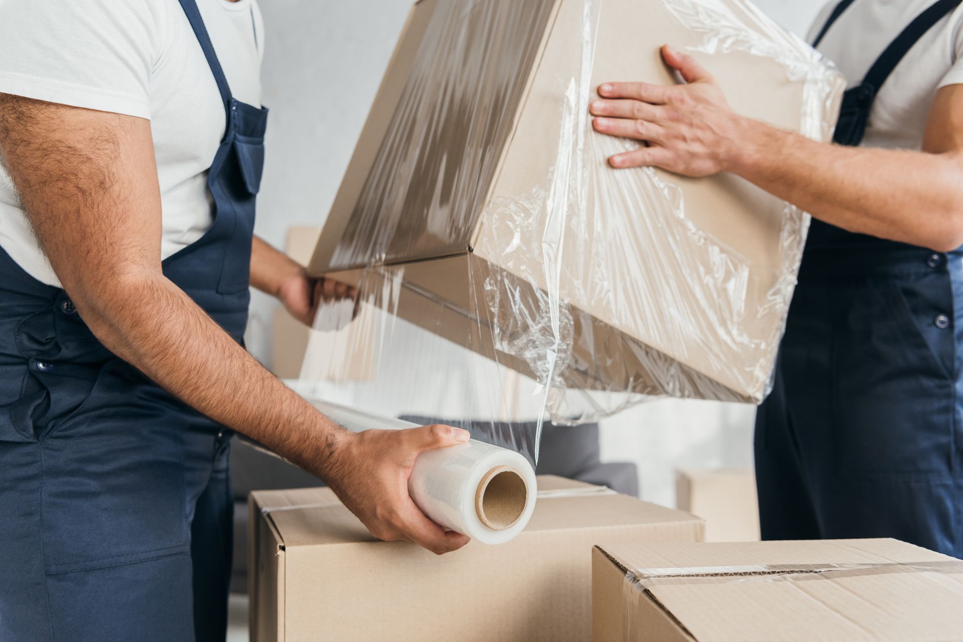 Two movers carrying a wrapped box and packing tape among moving boxes
