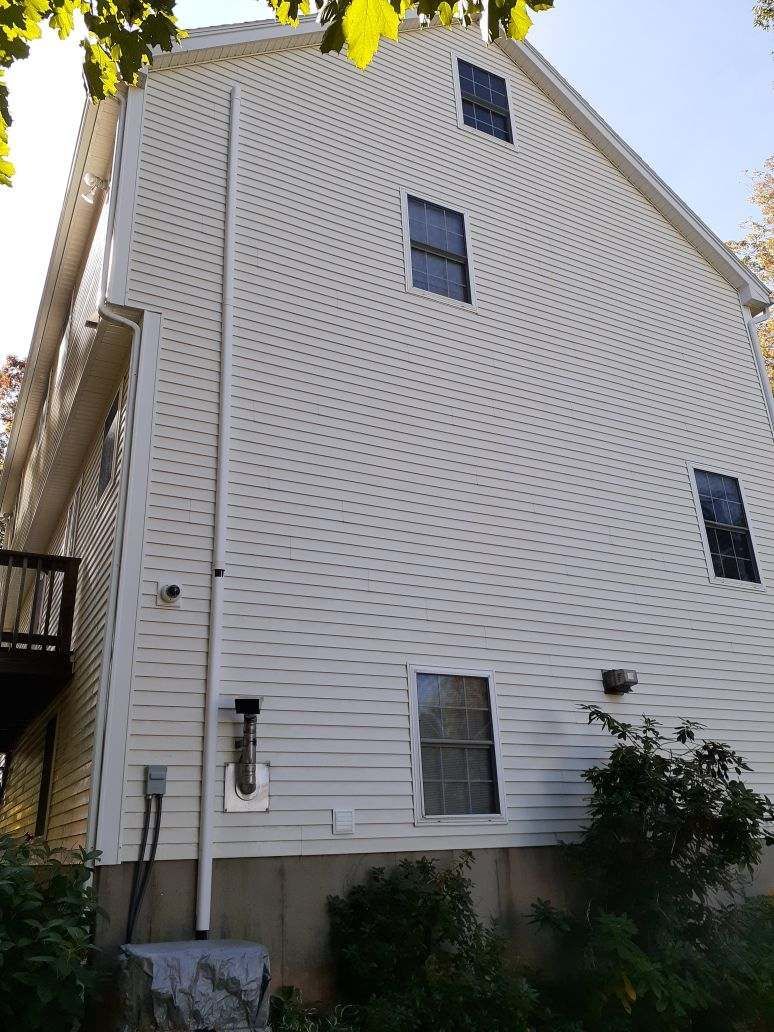 A white-sided house exterior with four windows, a pipe, and some greenery at the foundation.