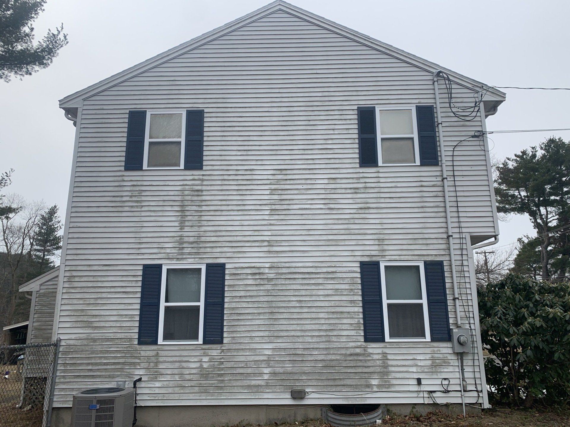 A two-story house exterior with weathered white siding, dark shutters, and four windows, set against a cloudy sky.