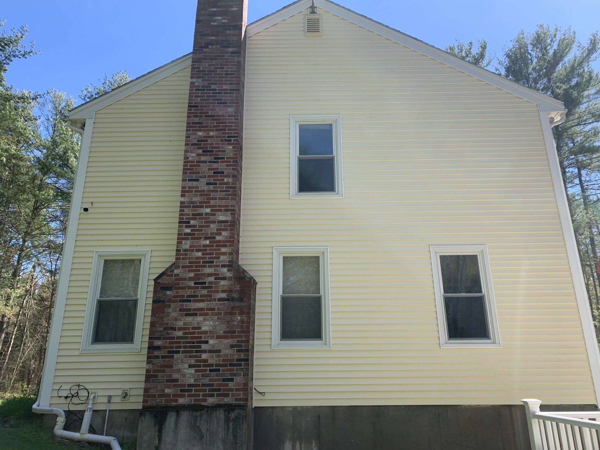 A yellow two-story house exterior with a prominent brick chimney and three windows, set against a sunny, wooded backdrop.