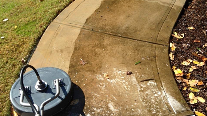 A surface cleaner is used to power wash a curved concrete walkway, showing a clear contrast between clean and dirty areas.