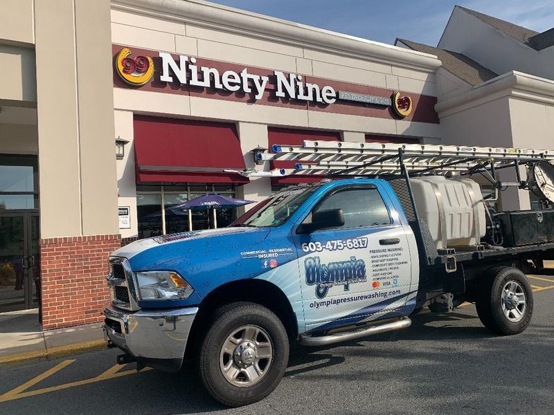 A blue Olympus service truck with a ladder rack and water tank parked in front of a Ninety Nine restaurant.