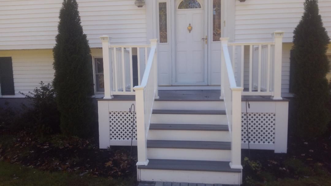 A front house entrance with gray wooden stairs, white railings, and white lattice skirting, framed by two tall evergreens.