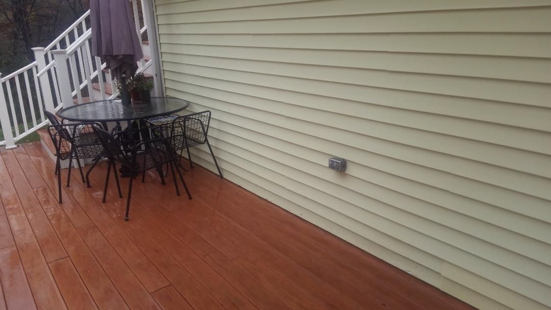 A patio set with a round table and chairs sits on a wooden deck next to a light yellow house with siding.