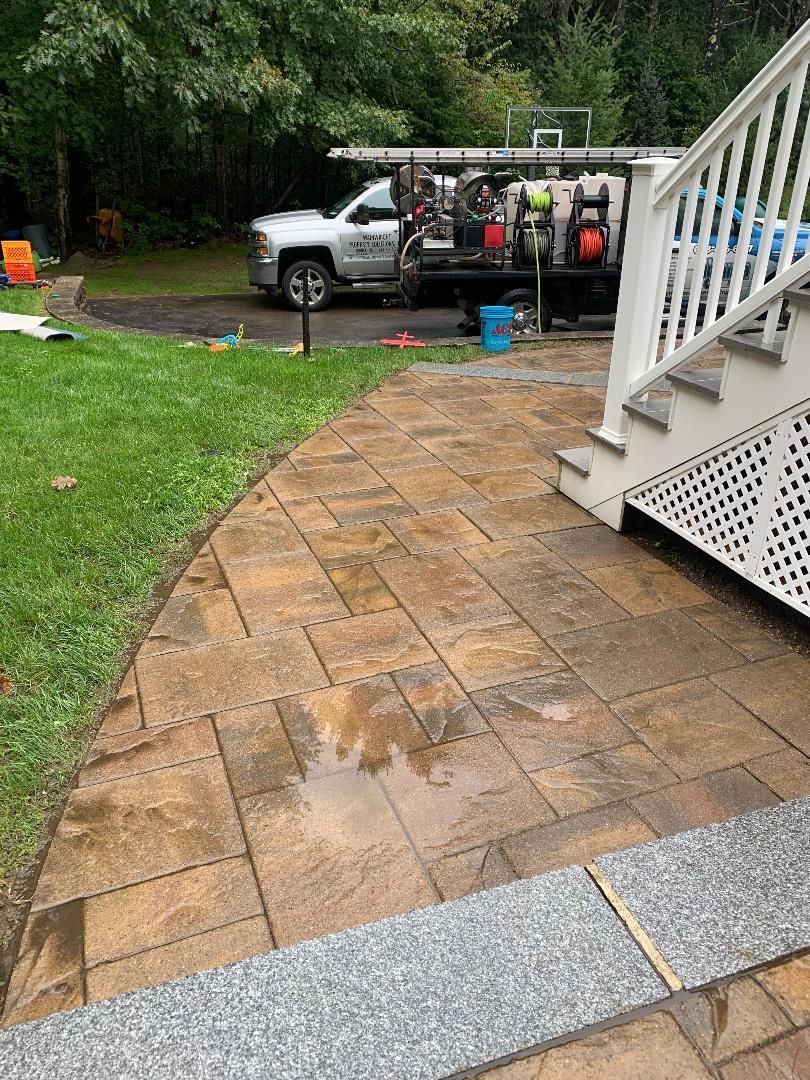 A newly installed tan paver patio leads to white deck stairs, with a work truck parked on a paved driveway in the background.