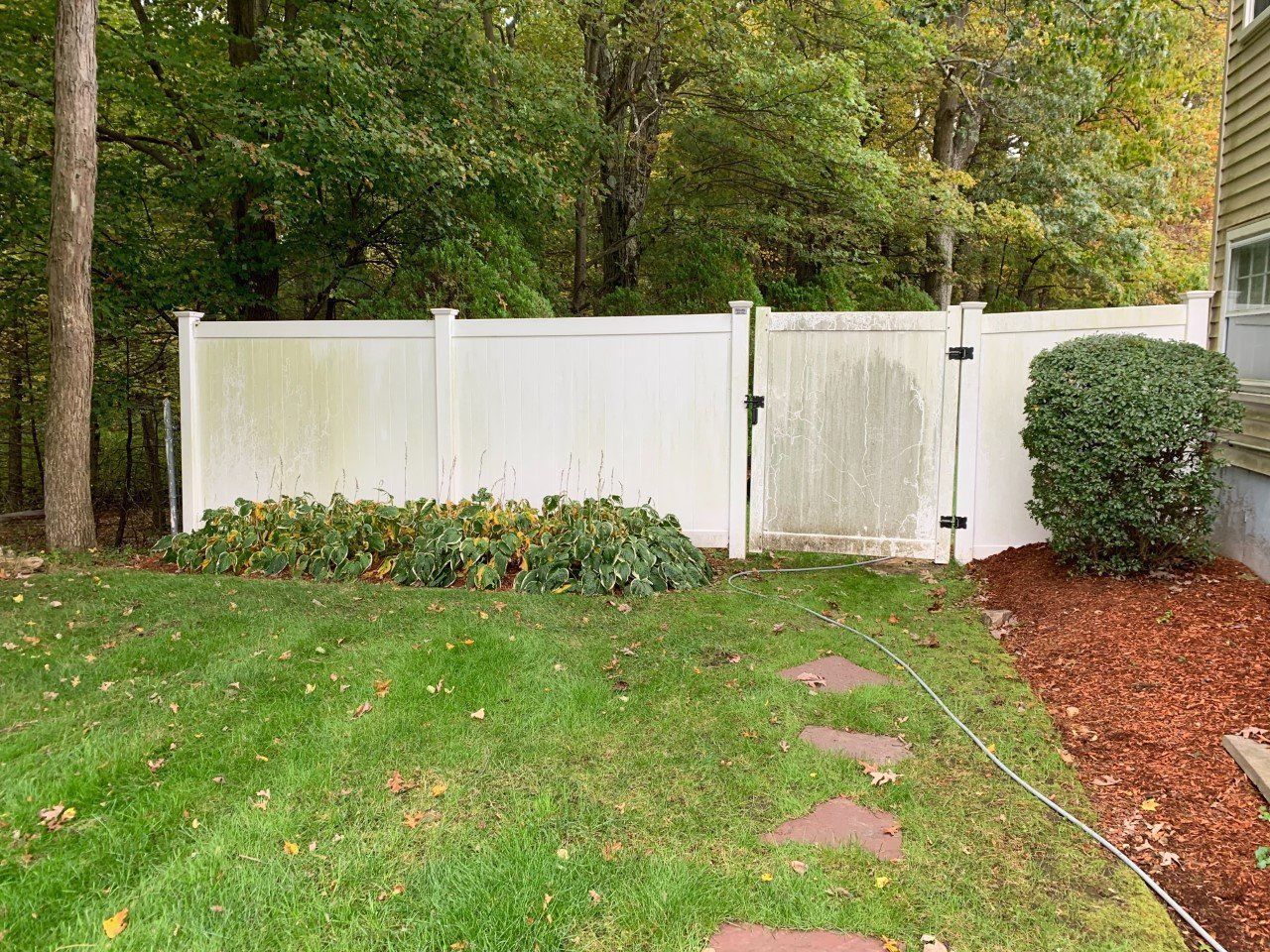 A white vinyl fence with a gate stands in a backyard, beside a green bush and a mulch bed, with trees in the background.