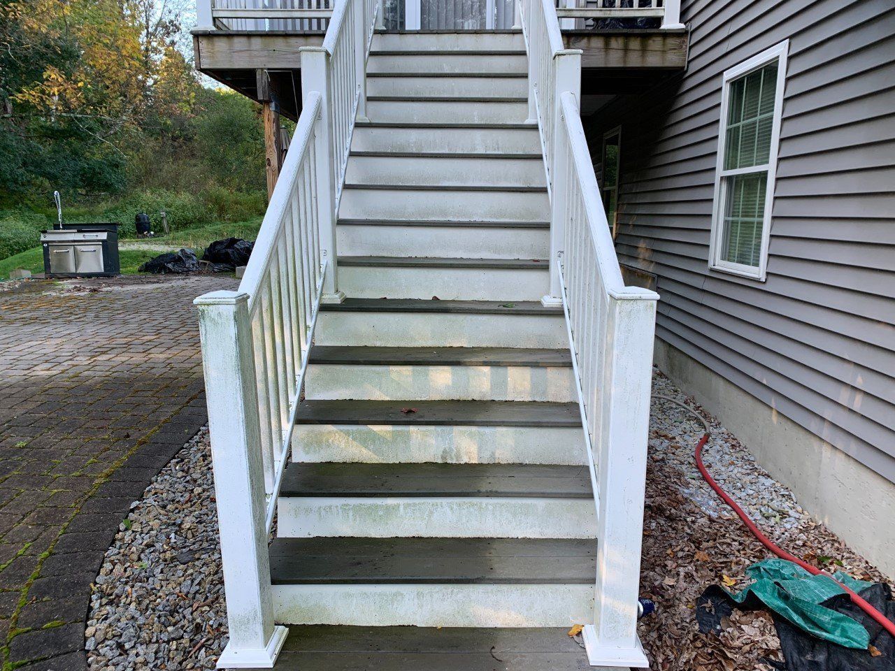 A set of wooden stairs with white railings leading up to a deck beside a house with grey siding and a window.