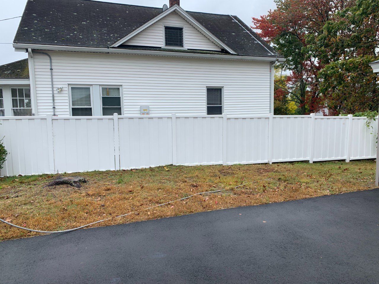A white vinyl fence stands in front of a white house with a dark roof and an asphalt driveway in the foreground.