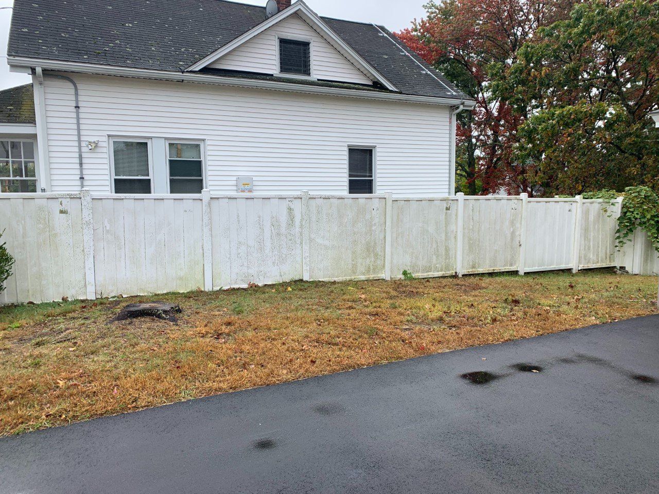 A white house with a dark shingled roof and vinyl siding stands behind a long, weathered white privacy fence.