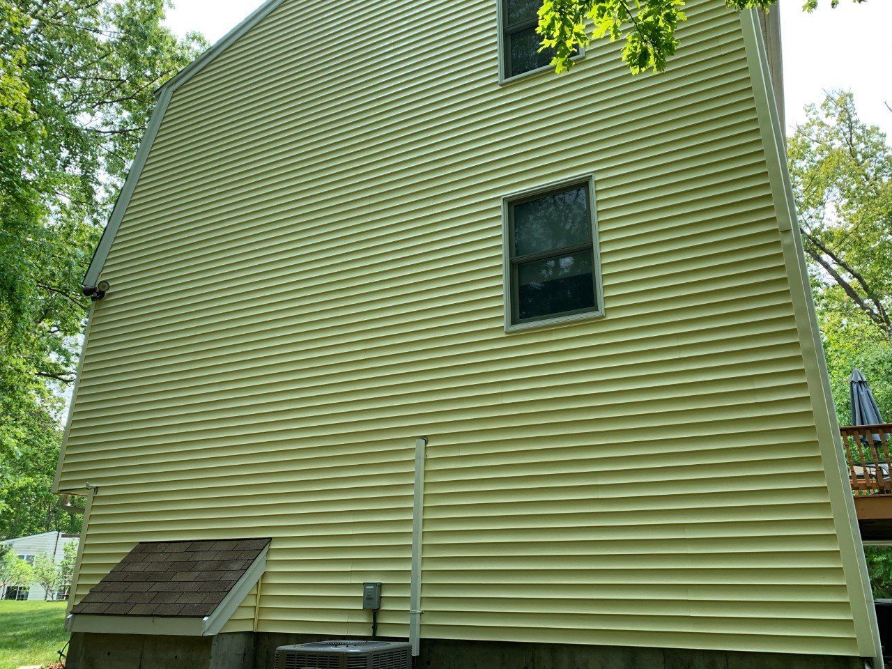 A yellow two-story house exterior with a prominent brick chimney and three windows, set against a sunny, wooded backdrop.