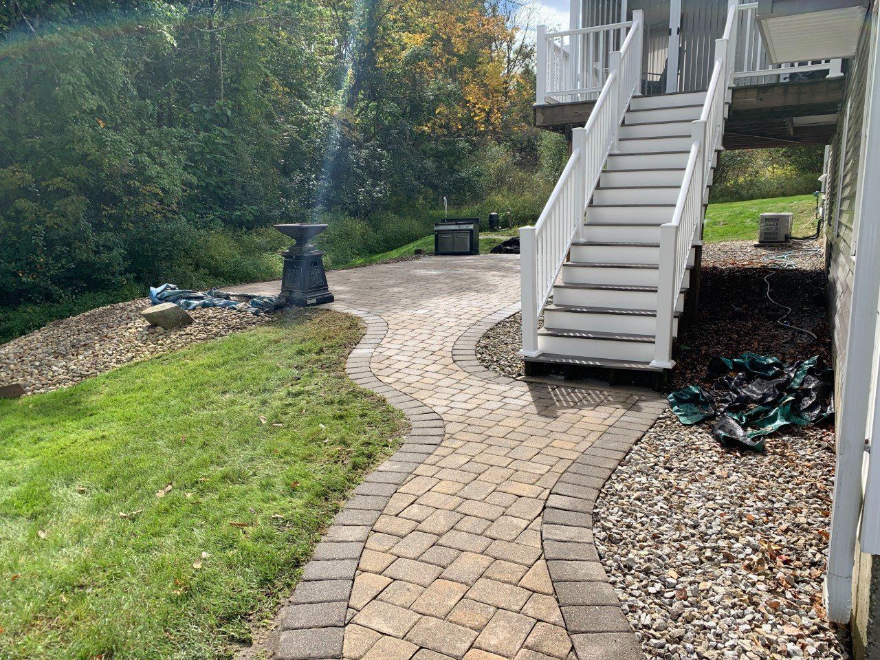 A tan stone paver walkway leads toward white stairs under a raised deck, surrounded by grass, gravel, and trees.