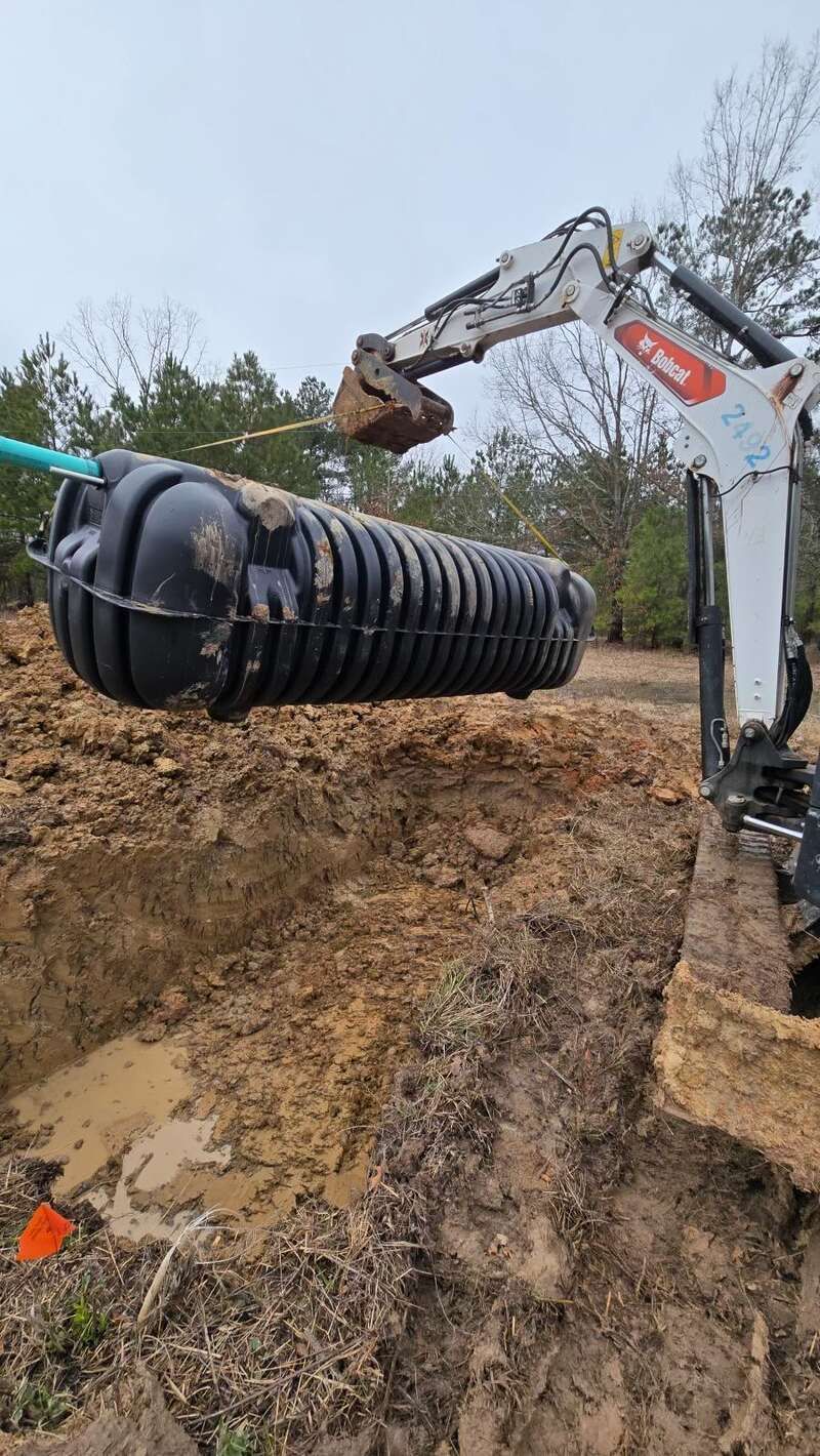 A Bobcat excavator lowers a black septic tank into a trench dug in a muddy, outdoor setting.